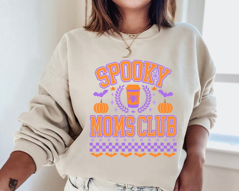 May include: A beige sweatshirt with a purple and orange graphic design that says "Spooky Moms Club". The graphic includes a coffee cup with a spider, pumpkins, bats, and a spiderweb.