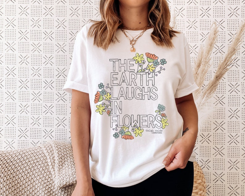 Pode incluir: Camiseta branca com um design floral e o texto "The Earth Laughs in Flowers - Ralph Waldo Emerson".