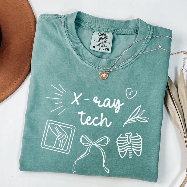 K&ouml;nnte beinhalten: Ein blaugr&uuml;nes T-Shirt mit dem Text "X-ray tech" in Wei&szlig;. Das Shirt zeigt verschiedene wei&szlig;e Illustrationen, darunter ein Herz, eine Schleife, einen Zweig, einen Brustkorb und ein R&ouml;ntgenbild eines Knies.
