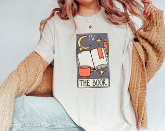 May include: A white t-shirt with a graphic of a tarot card. The card is purple and features a crescent moon, stars, a book, a cup of coffee, and the text "The Book".