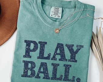 May include: A seafoam green t-shirt with the words "PLAY BALL." in navy blue block letters. A silver necklace with a small pendant rests on the shirt. The shirt is folded, and a brown hat is visible in the corner.