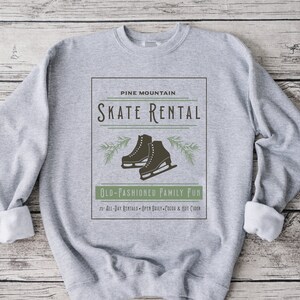 K&ouml;nnte beinhalten: Grauer Pullover mit einem Vintage-Grafikdesign mit einem Paar Schlittschuhen, Kiefern&auml;sten und dem Text "PINE MOUNTAIN SKATE RENTAL OLD-FASHIONED FAMILY FUN 25 ALL-DAY RENTALS OPEN DAILY COCOA & HOT CIDER".