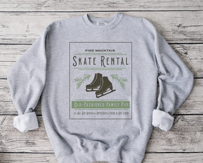 K&ouml;nnte beinhalten: Grauer Pullover mit einem Vintage-Grafikdesign mit einem Paar Schlittschuhen, Kiefern&auml;sten und dem Text "PINE MOUNTAIN SKATE RENTAL OLD-FASHIONED FAMILY FUN 25 ALL-DAY RENTALS OPEN DAILY COCOA & HOT CIDER".