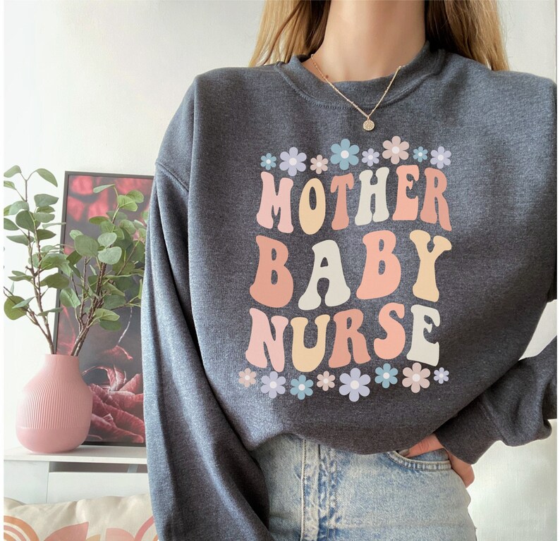 May include: A dark gray sweatshirt with the text "MOTHER BABY NURSE" in colorful retro-style lettering. The text is surrounded by a row of small pastel flowers.