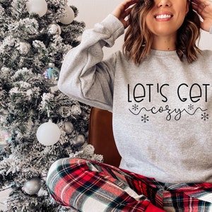 May include: A gray sweatshirt with the text "Let's Get Cozy" in black lettering with snowflake accents. The person wearing the sweatshirt is sitting in front of a Christmas tree and wearing red and black plaid pajama pants.
