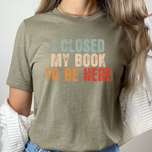 Puede incluir: Una camiseta verde con el texto "I CLOSED MY BOOK TO BE HERE" en una fuente retro con colores azul, naranja y rojo.
