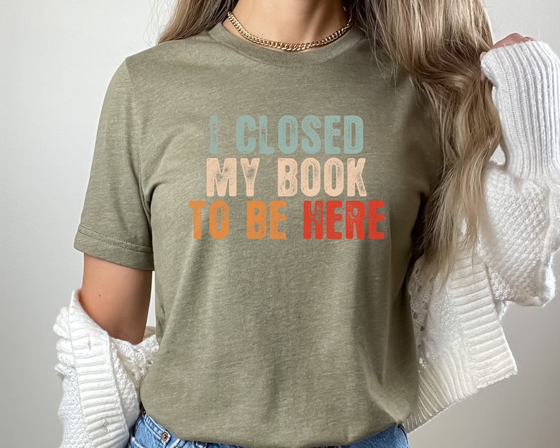 Puede incluir: Una camiseta verde con el texto "I CLOSED MY BOOK TO BE HERE" en una fuente retro con colores azul, naranja y rojo.