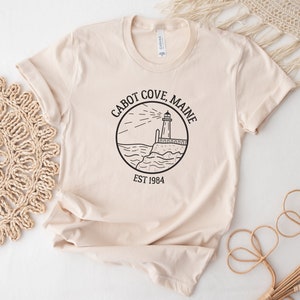 Puede incluir: Una camiseta beige con un gr&aacute;fico negro de un faro y el texto "Cabot Cove, Maine Est 1984".