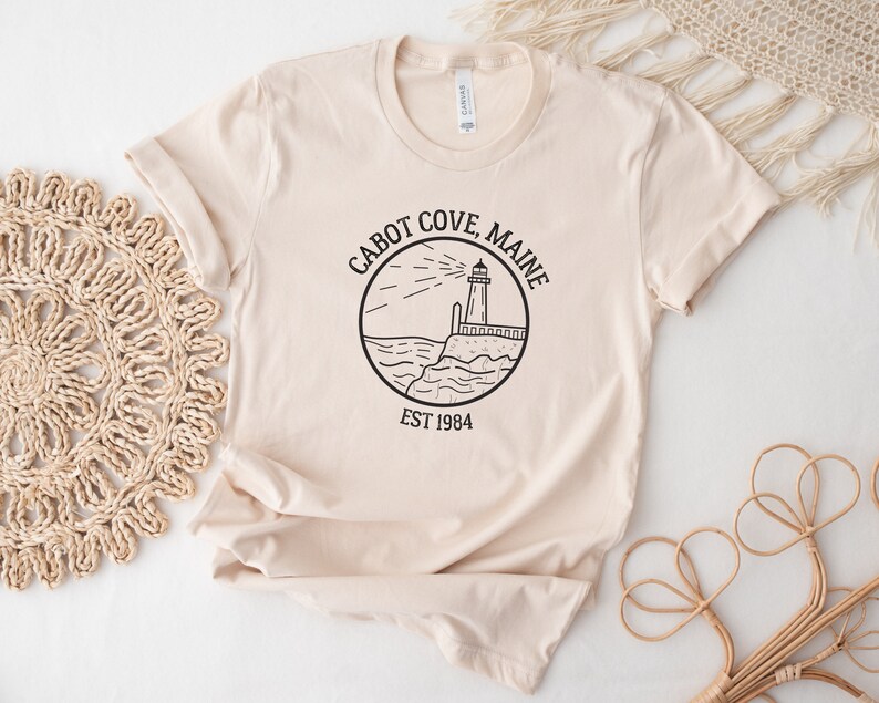 Puede incluir: Una camiseta beige con un gr&aacute;fico negro de un faro y el texto "Cabot Cove, Maine Est 1984".