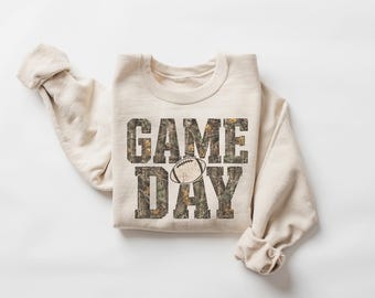 May include: A cream-colored crewneck sweatshirt featuring the text GAME DAY in a block font with a camouflage pattern. A small, simple line drawing of an American football is positioned in the center of the text between the words GAME and DAY.