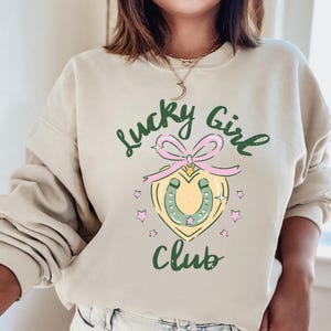 May include: A beige sweatshirt with the text "Lucky Girl Club" in green and pink lettering. A green horseshoe with a pink bow is in the center of the design.