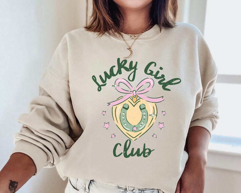May include: A beige sweatshirt with the text "Lucky Girl Club" in green and pink lettering. A green horseshoe with a pink bow is in the center of the design.