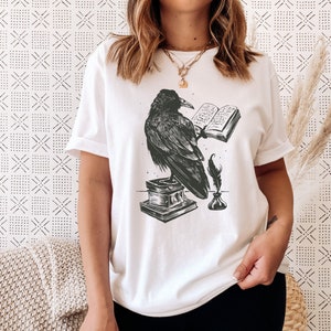 Puede incluir: Camiseta blanca con una ilustraci&oacute;n en blanco y negro de un cuervo sosteniendo un libro abierto, una pluma y un tintero.