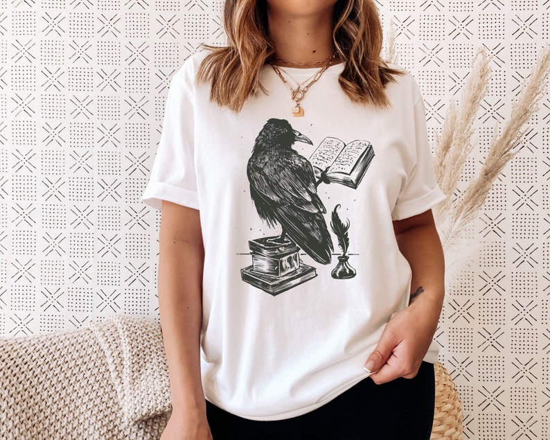 Puede incluir: Camiseta blanca con una ilustraci&oacute;n en blanco y negro de un cuervo sosteniendo un libro abierto, una pluma y un tintero.
