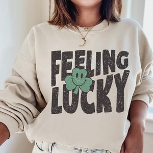 May include: A beige sweatshirt with the text "Feeling Lucky" in black with a green shamrock with a smiley face.