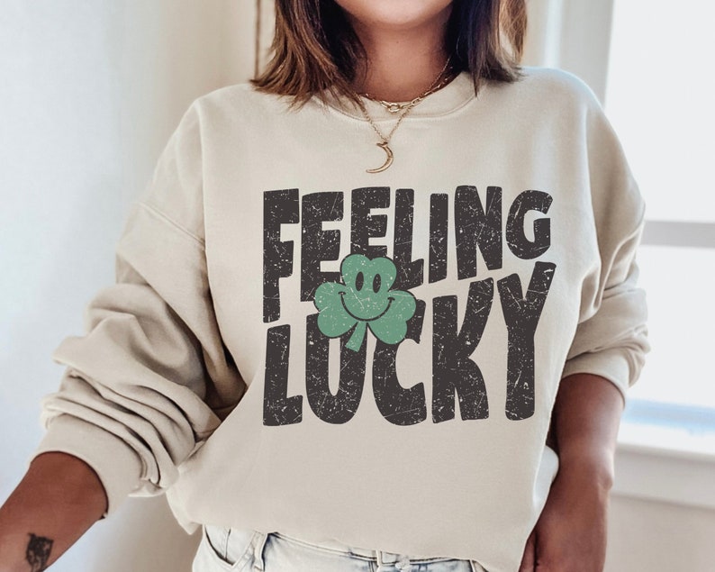 May include: A beige sweatshirt with the text "Feeling Lucky" in black with a green shamrock with a smiley face.