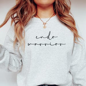 May include: A light gray sweatshirt with the words "endo warrior" in black script. The sweatshirt is a crew neck style and appears to be made of a soft material. The overall aesthetic is casual and comfortable.