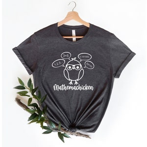 May include: A dark gray t-shirt with a white graphic of a cartoon chicken wearing glasses and thinking about math equations. The text "Mathemachicken" is printed below the chicken.