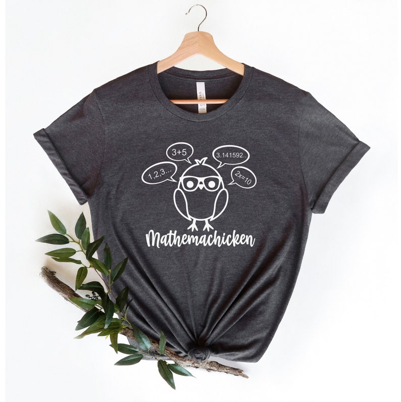 May include: A dark gray t-shirt with a white graphic of a cartoon chicken wearing glasses and thinking about math equations. The text "Mathemachicken" is printed below the chicken.