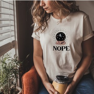 May include: White t-shirt with a black and white illustration of a snow globe with stars and a crescent moon. The text "NOPE." is printed below the snow globe.