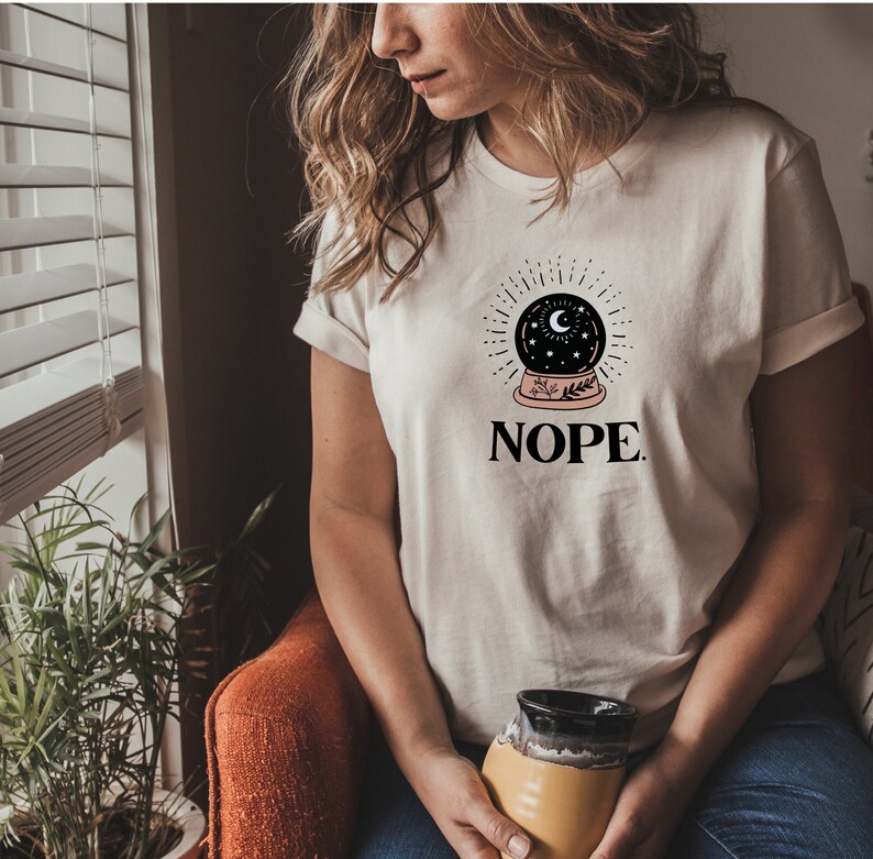 May include: White t-shirt with a black and white illustration of a snow globe with stars and a crescent moon. The text "NOPE." is printed below the snow globe.