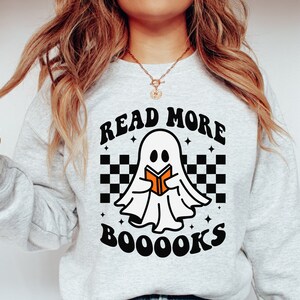 May include: A gray sweatshirt with a white ghost holding a book and the text "Read More Booooks" in black with a checkered pattern behind the ghost.