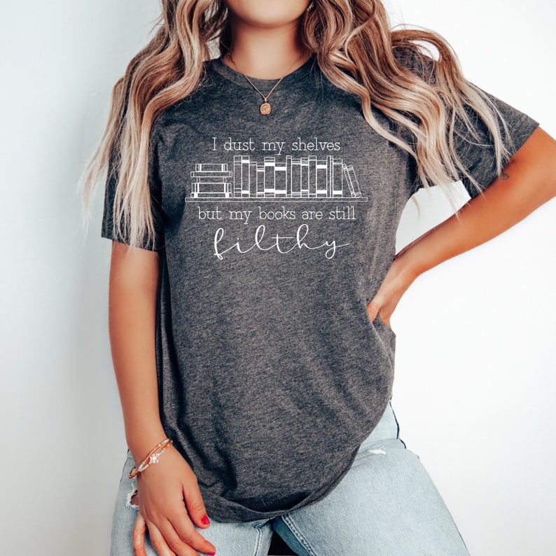 Peut inclure: Un t-shirt gris fonc&eacute; avec un motif blanc qui dit "I dust my shelves but my books are still filthy" avec un dessin de livres sur une &eacute;tag&egrave;re.