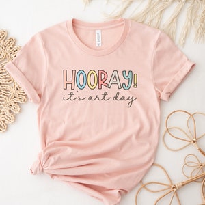 May include: A pink t-shirt with the text "HOORAY! it's art day" in colourful letters.