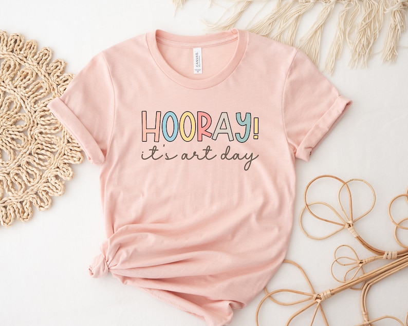 May include: A pink t-shirt with the text "HOORAY! it's art day" in colourful letters.