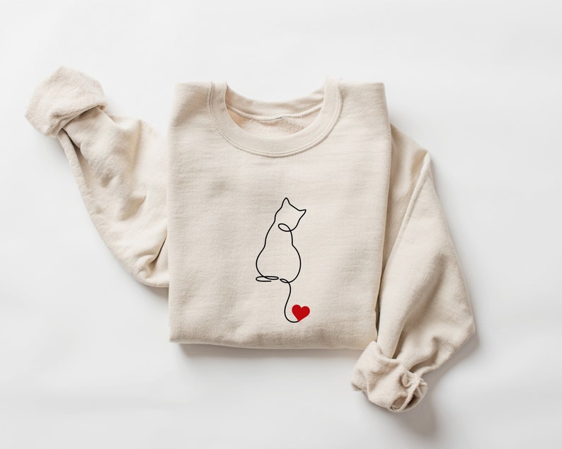 Peut inclure: Un sweat-shirt beige avec un dessin au trait noir d'un chat avec un c&oelig;ur rouge sur sa queue.