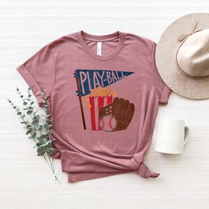 May include: A mauve t-shirt with a graphic of a baseball glove, popcorn, and a pennant that says "PLAY-BALL".