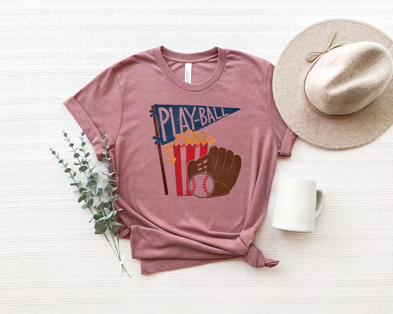 May include: A mauve t-shirt with a graphic of a baseball glove, popcorn, and a pennant that says "PLAY-BALL".