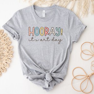 May include: Grey t-shirt with the text "HOORAY! it's art day" in colourful letters.