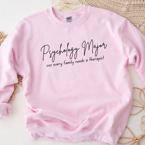以下が含まれることがあります： ピンクのトレーナーに「Psychology Major cuz every family needs a therapist」という黒い文字が書かれています。