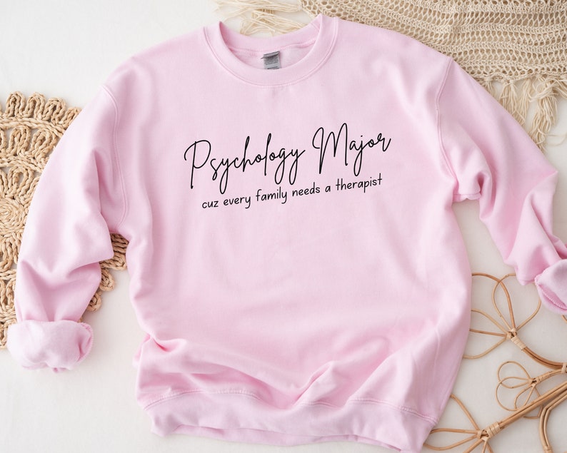 以下が含まれることがあります： ピンクのトレーナーに「Psychology Major cuz every family needs a therapist」という黒い文字が書かれています。