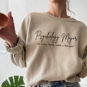 以下が含まれることがあります： 前面に「Psychology Major cuz every family needs a therapist」という文字がプリントされたベージュのトレーナーです。