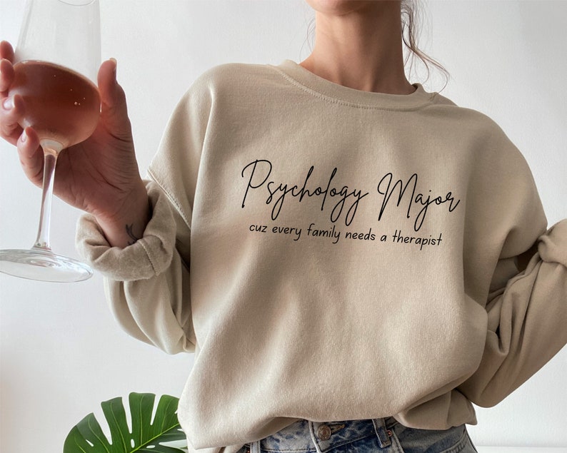 以下が含まれることがあります： 前面に「Psychology Major cuz every family needs a therapist」という文字がプリントされたベージュのトレーナーです。
