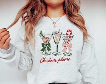 May include: A light gray crewneck sweatshirt featuring a graphic of three cocktail glasses decorated with a green bow, a red candy cane, and a sprig of rosemary. The text "Christmas, please" is printed in red cursive script below the glasses.