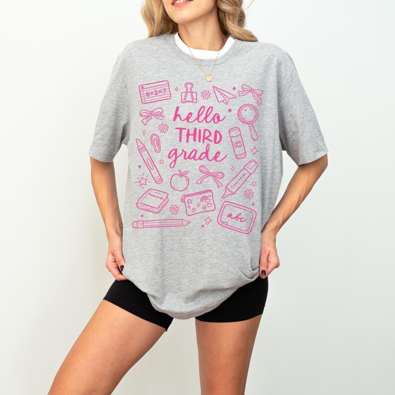 May include: A gray t-shirt with a pink graphic that says "hello THIRD grade" surrounded by school-themed illustrations. The illustrations include a pencil, apple, and a magnifying glass.