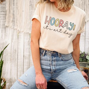 May include: A light beige t-shirt with the text "HOORAY! it's art day" in colourful letters.