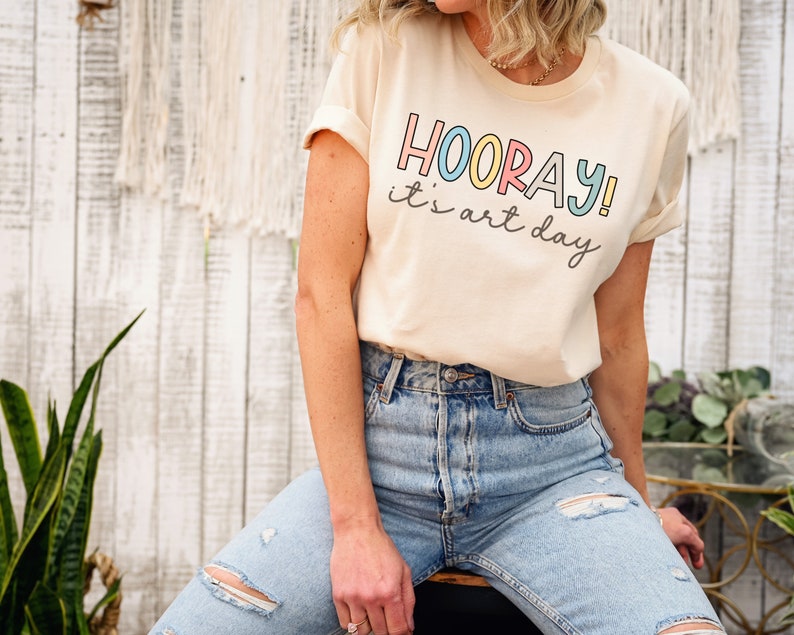 May include: A light beige t-shirt with the text "HOORAY! it's art day" in colourful letters.