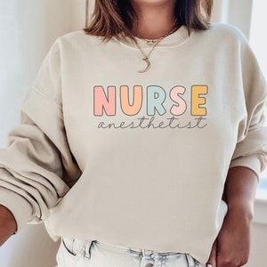 May include: A light beige sweatshirt with the text "NURSE anesthetist" in colorful letters. The letters are outlined in black and have a pastel color scheme.