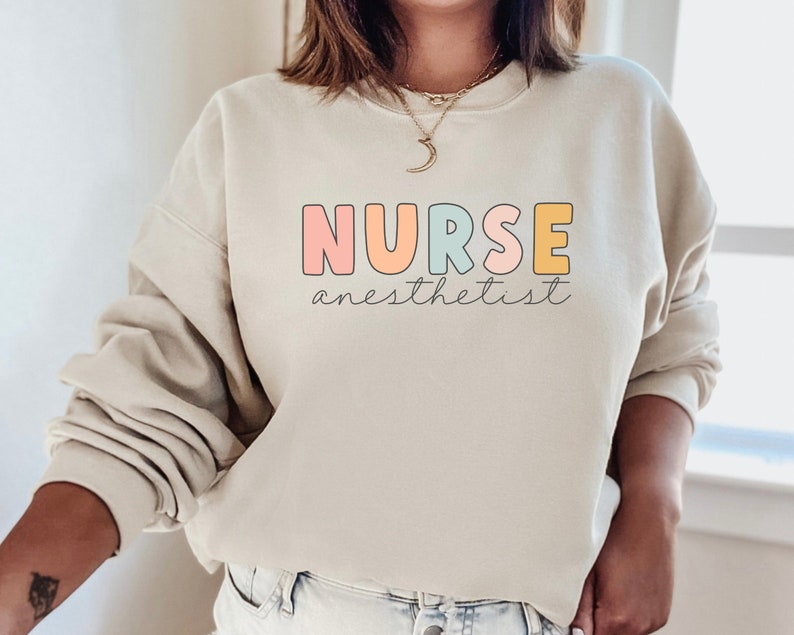 May include: A light beige sweatshirt with the text "NURSE anesthetist" in colorful letters. The letters are outlined in black and have a pastel color scheme.