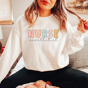 May include: White sweatshirt with the text "NURSE anesthetist" in colorful letters. The letters are outlined in black.