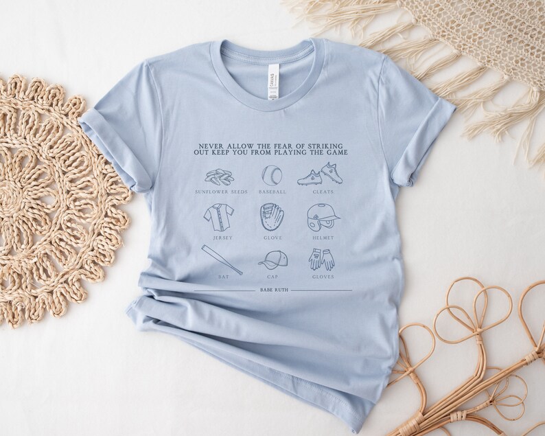 Peut inclure: T-shirt bleu clair avec un motif sur le th&egrave;me du baseball. Le motif comprend le texte "NEVER ALLOW THE FEAR OF STRIKING OUT KEEP YOU FROM PLAYING THE GAME" et des illustrations d'&eacute;quipements de baseball, notamment une batte, une balle, un gant, des crampons, un casque et une casquette.