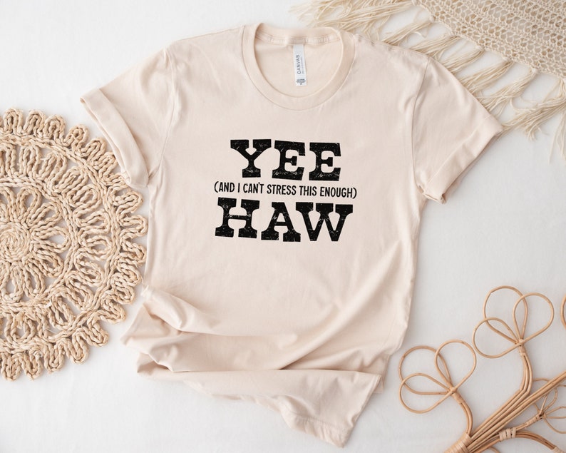 Op de afbeelding: Een beige T-shirt met de tekst "YEE (AND I CAN'T STRESS THIS ENOUGH) HAW" in zwart.