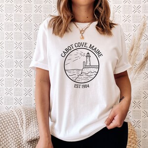 Puede incluir: Camiseta blanca con un gr&aacute;fico negro de un faro, olas y el texto "CABOT COVE, MAINE EST 1984".