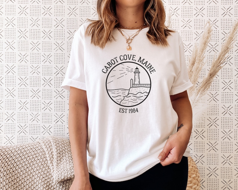 Puede incluir: Camiseta blanca con un gr&aacute;fico negro de un faro, olas y el texto "CABOT COVE, MAINE EST 1984".