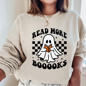 May include: A cream-colored sweatshirt with a black and white checkered pattern, a ghost holding a book, and the text "Read More Booooks".