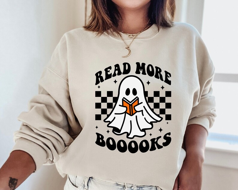 May include: A cream-colored sweatshirt with a black and white checkered pattern, a ghost holding a book, and the text "Read More Booooks".